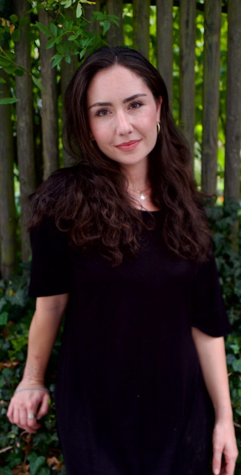 Giorgia Nebuloni Heilpraktikerin für Psychotherapie Das Bild zeigt Giorgia Nebuloni, eine Heilpraktikerin für Psychotherapie aus Köln, vor einem Holzzaun, der von üppigem, grünem Laub und Pflanzen umgeben ist. Sie trägt ein schwarzes Kleid und lächelt sanft. Giorgia hat lange, dunkle Haare und trägt goldene Ohrringe. Die natürliche und beruhigende Umgebung reflektiert ihre einfühlsame und harmonische Herangehensweise in der Psychotherapie.
